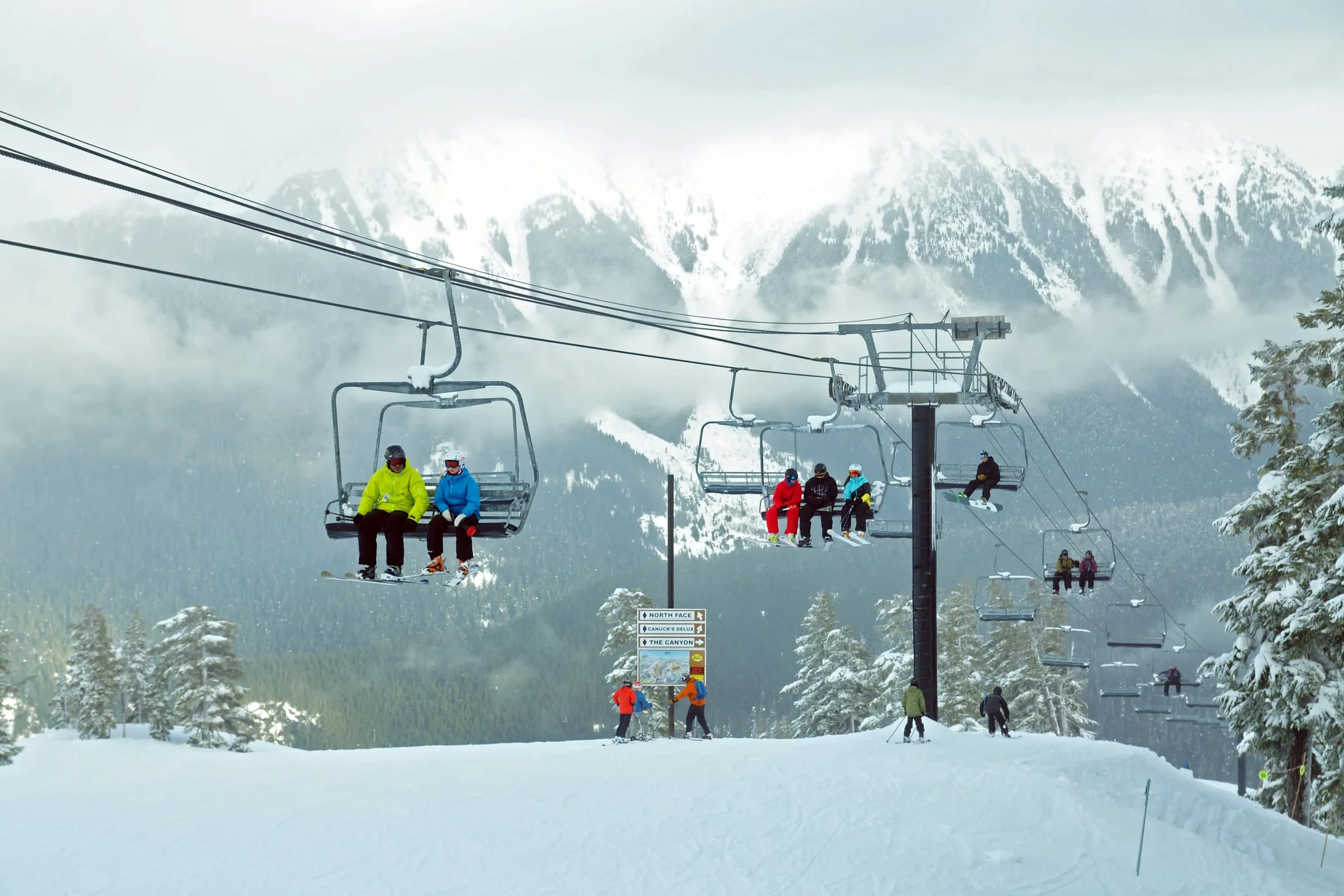 Riders on a Mt. Baker chairlift above a trail sign for North Face, Canuck's Delight and The Canyon