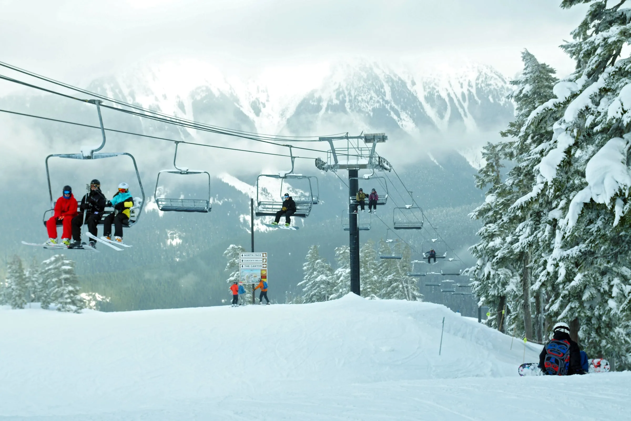 Skiers and snowboarders riding a chairlift at Mt. Baker Ski Area past a trail sign for North Face, Canuck's Deluxe and The Canyon