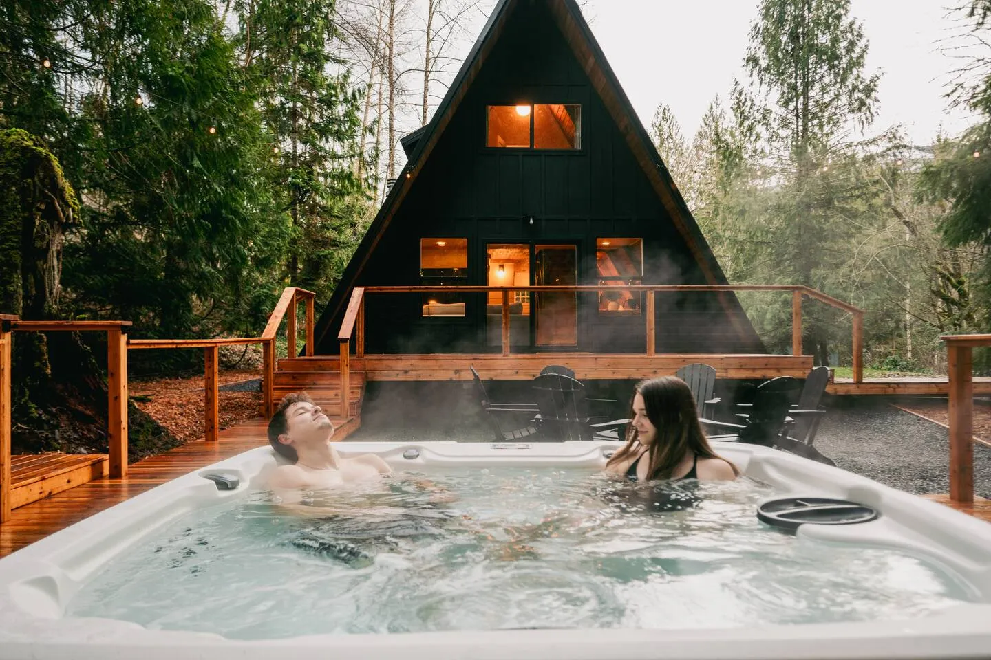 Couple soaking in the steaming hot tub in front of the black A-frame cabin